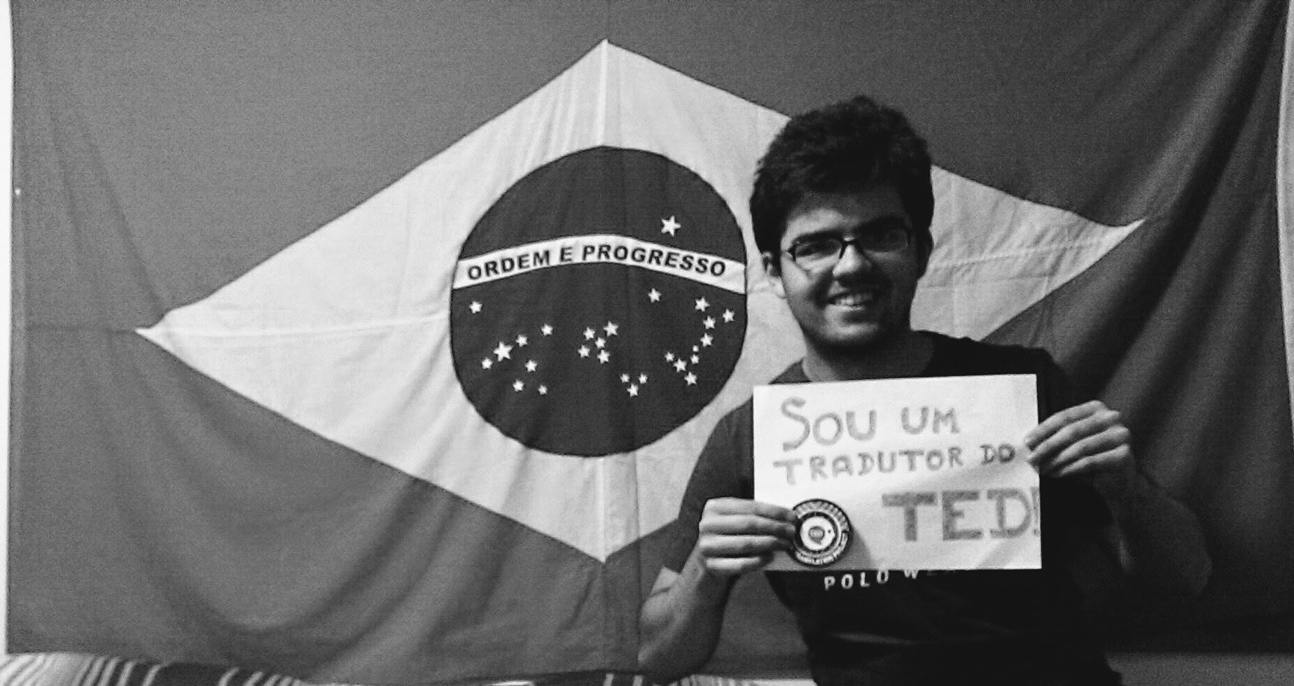 Picture of the author smiling and sitting in front of a Brazil flag holding a sign saying "I'm a TED Translator"