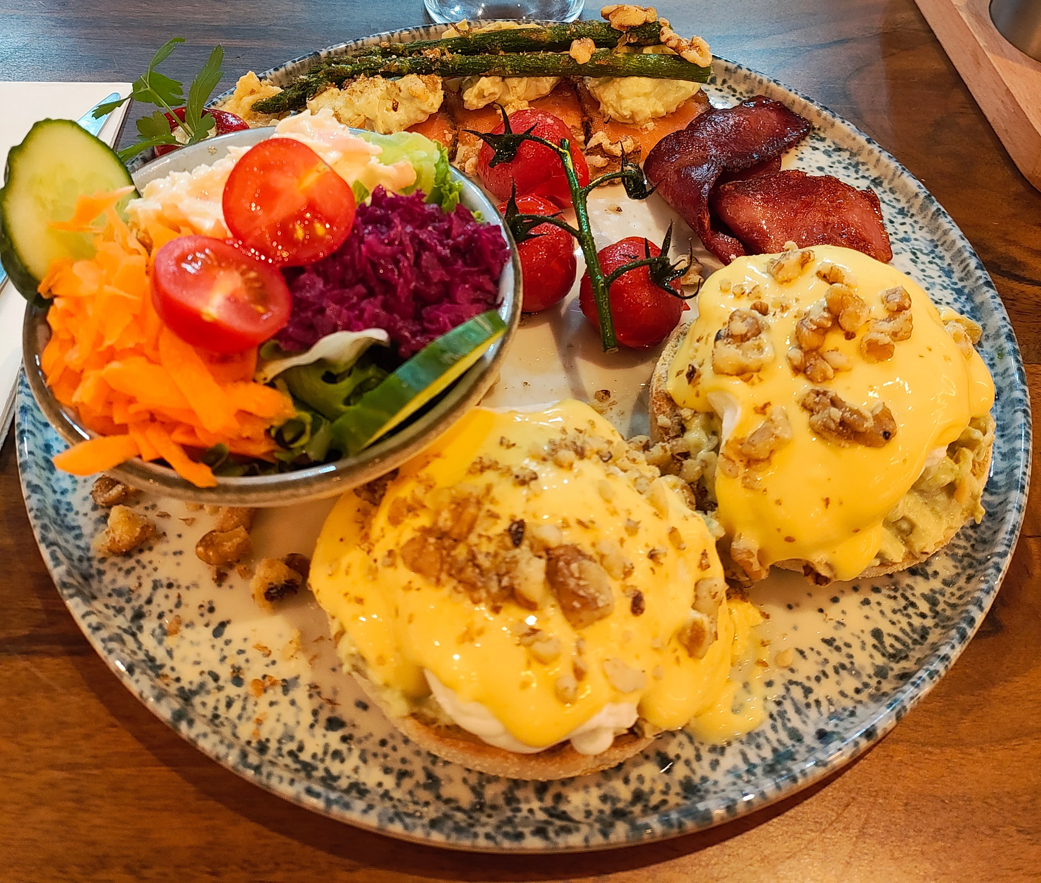 A plate with a bowl of salad, two buns with eggs and filling, a few tomatoes, asparagus, bacon and smoked salmon